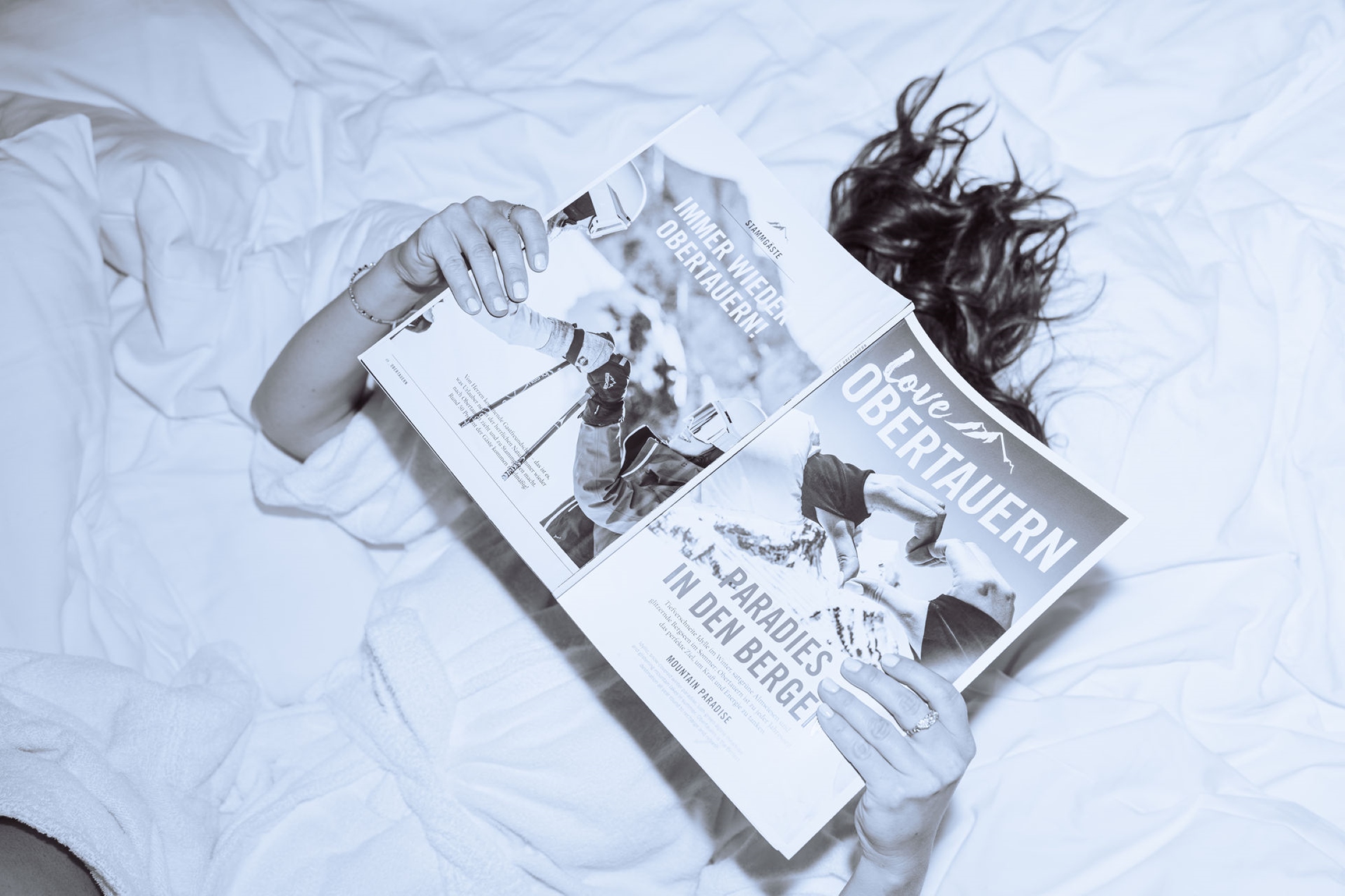 Black-and-white aerial view of a guest relaxing in bed at the Wagner Hotels in Obertauern, reading a magazine after a day in the snow.